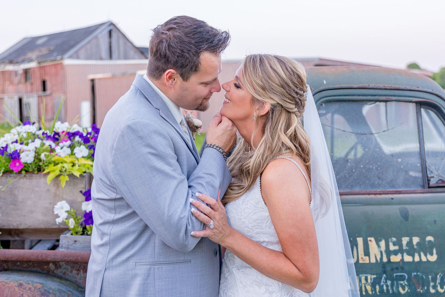 A tender moment between newlyweds near vintage truck with purple flowers in background.