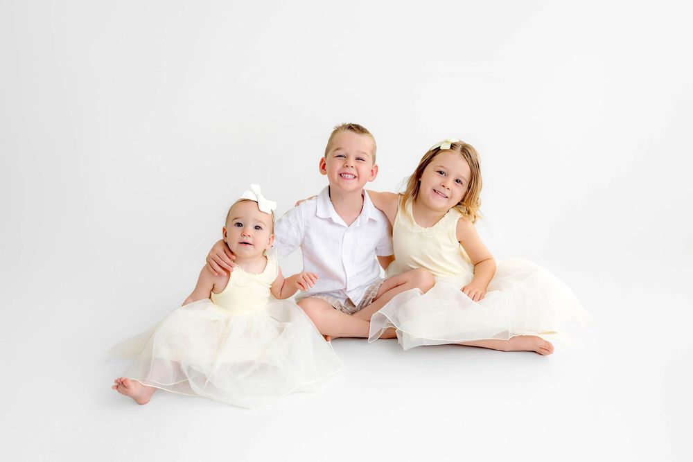 Three young siblings in white outfits pose together on a white background, smiling for a professional photo.