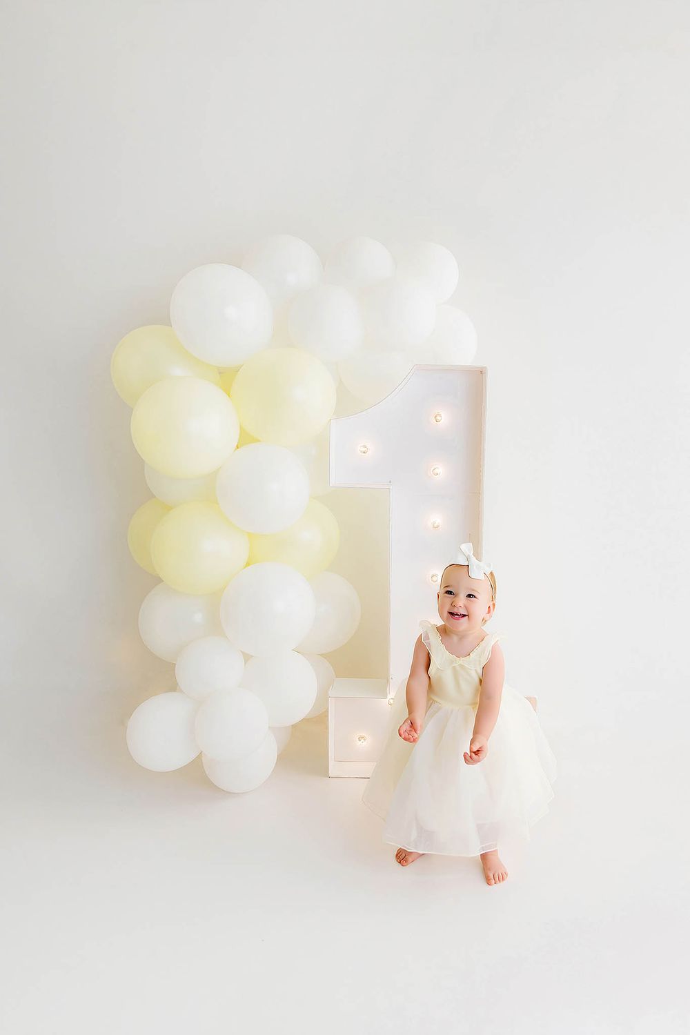 Toddler girl in white tulle dress standing by illuminated number 1 with yellow and white balloon arch.