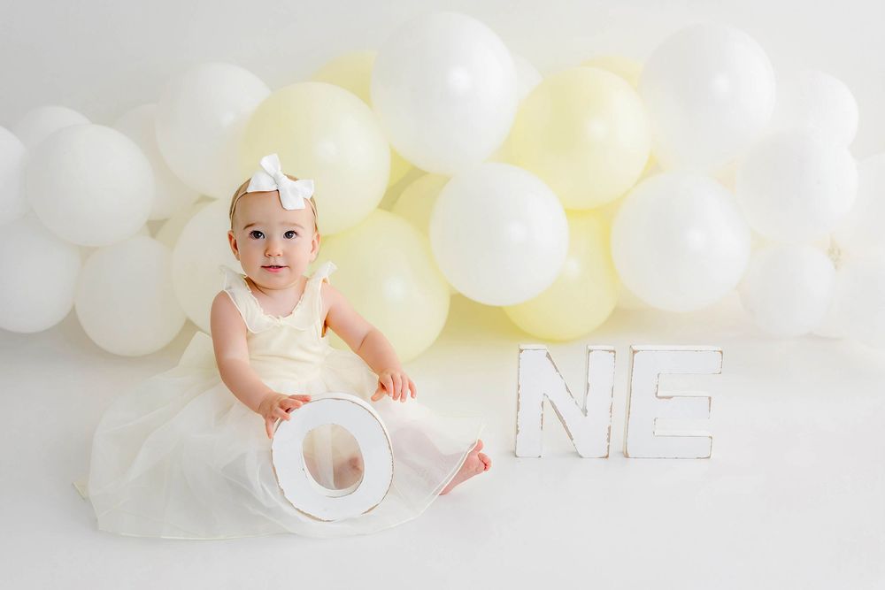 Adorable baby girl in cream tutu dress poses with ONE letters and white balloons at first birthday photo shoot.