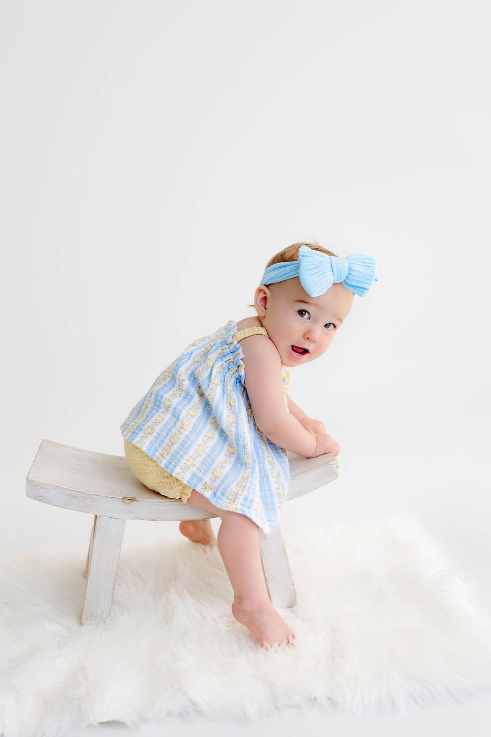 Baby girl in blue striped romper and bow headband standing at a small wooden bench on a white fur rug.