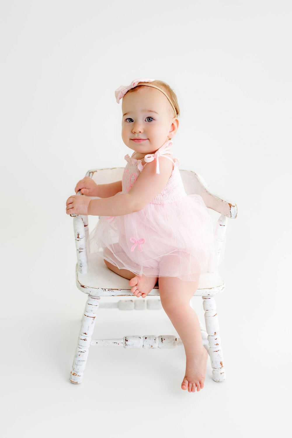 Adorable baby girl in pink tutu dress with bow headband sitting in a white vintage chair.