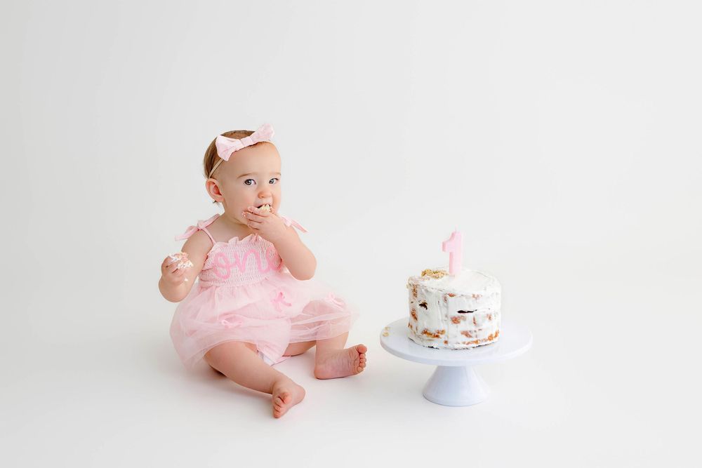 Baby girl in pink tutu sits beside first birthday cake, tasting frosting during cake smash photo session.