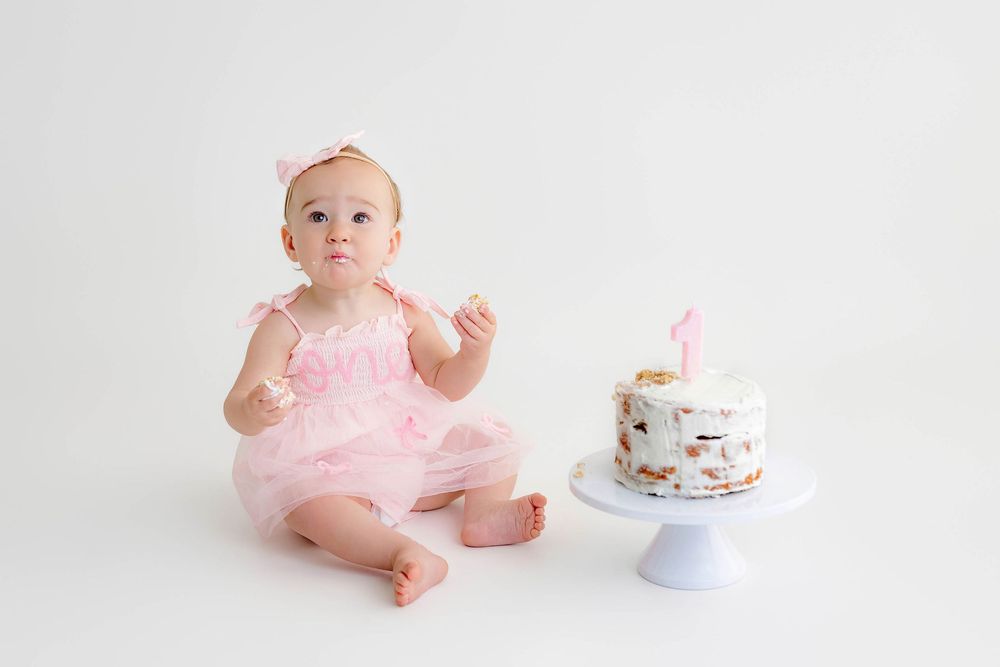Baby girl in pink tutu sits beside her first birthday cake with a number one candle on white background.