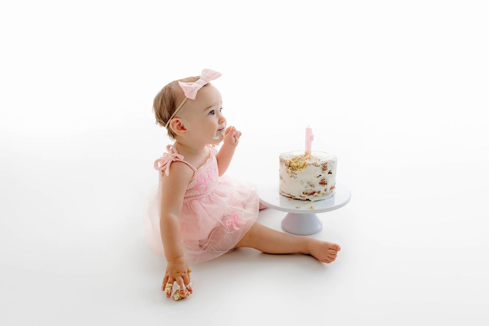 Baby girl in pink tutu romper reaching for her first birthday smash cake on a white cake stand.