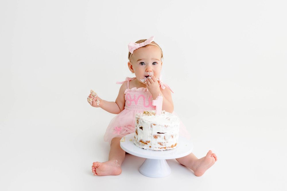 Cute baby girl in pink tutu dress eating her first birthday smash cake on a white cake stand.