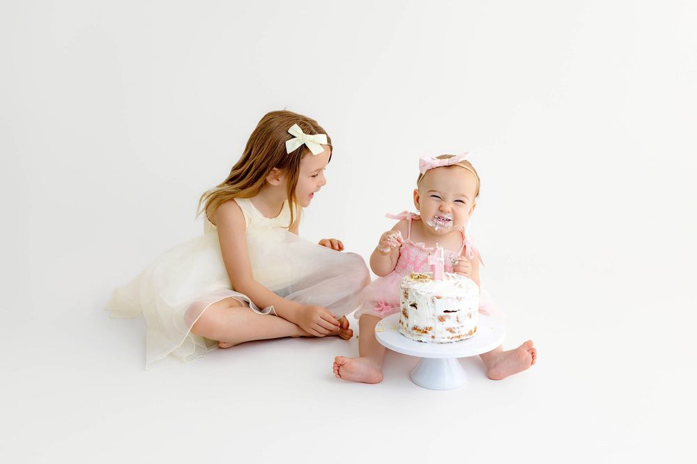 Two young sisters celebrate a first birthday cake smash in white studio, older girl watching baby sister in pink tutu.