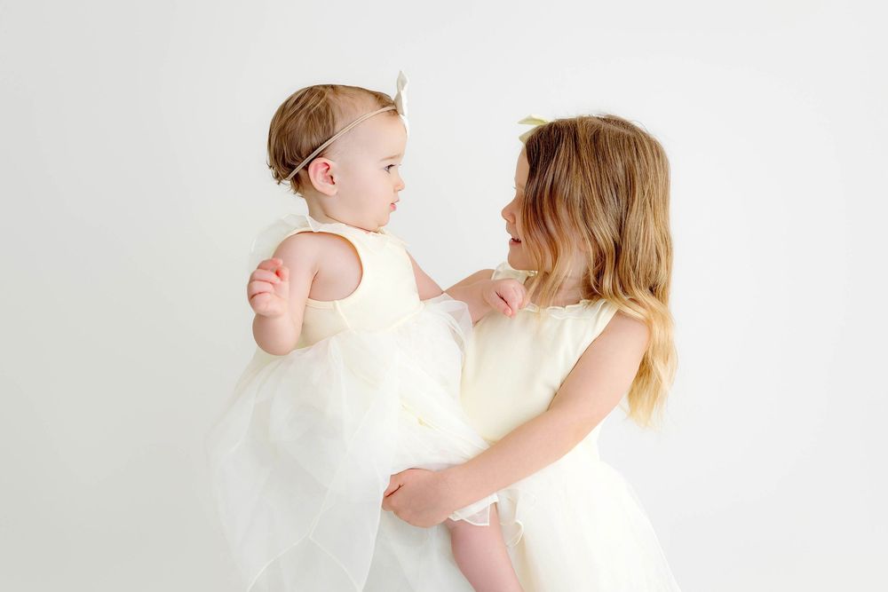 An older girl in a white dress lovingly holds a baby girl in a matching white tulle dress.