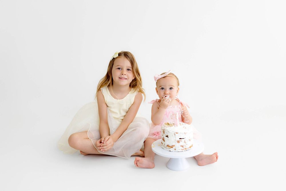 Two sisters pose together, older girl in white dress beside baby in pink eating first birthday cake.