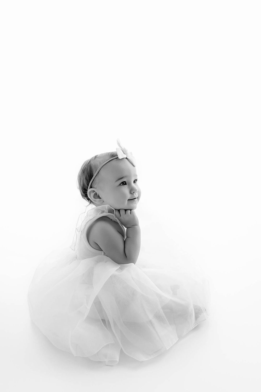 Black and white photo of an adorable baby girl in a tutu dress and bow headband sitting and looking up sweetly.