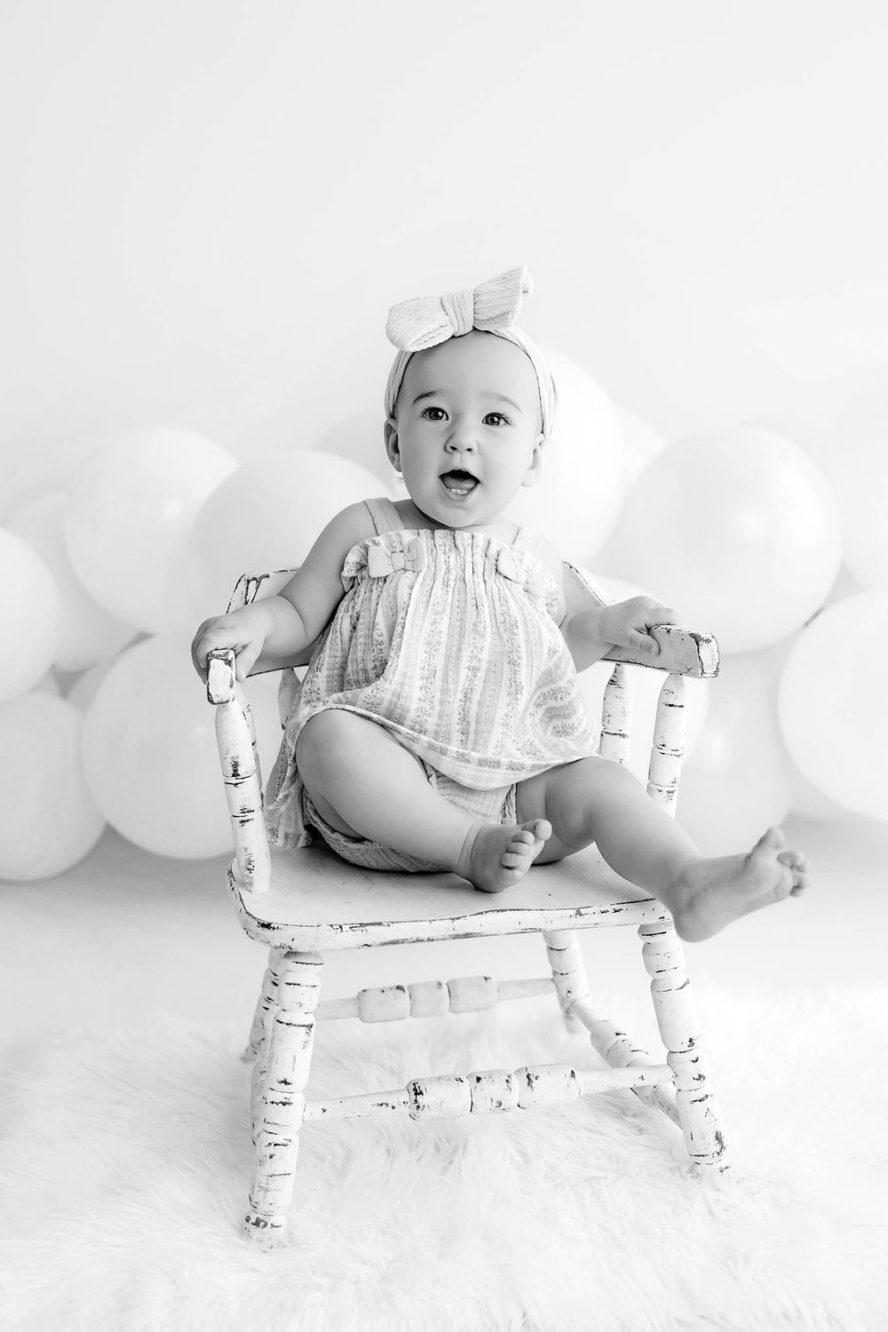Black and white photo of a cute baby girl sitting in a rustic white chair with balloons in the background.
