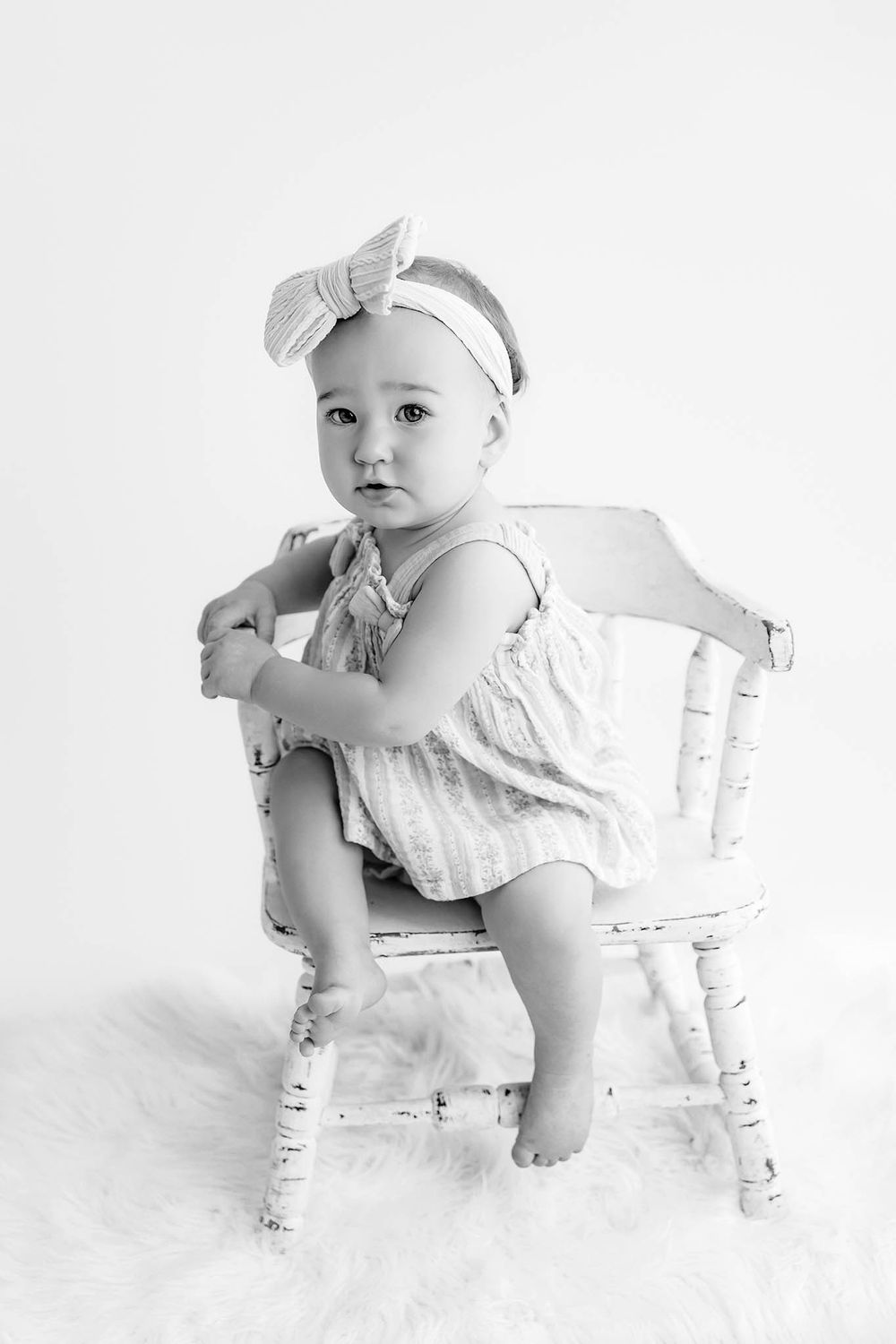 Black and white photo of a baby girl in a romper and bow headband sitting on a small white wooden chair.