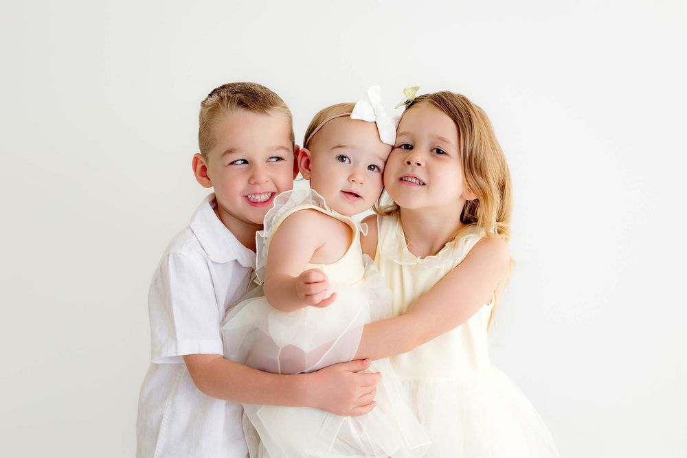 Two older siblings lovingly embrace a baby girl in white outfits against a white background.