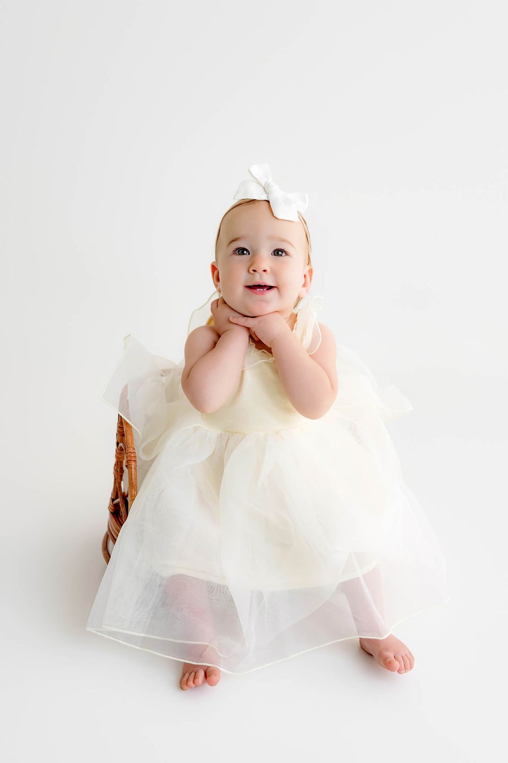 Adorable baby girl in cream tulle dress and white bow headband sitting in wicker basket on white background.
