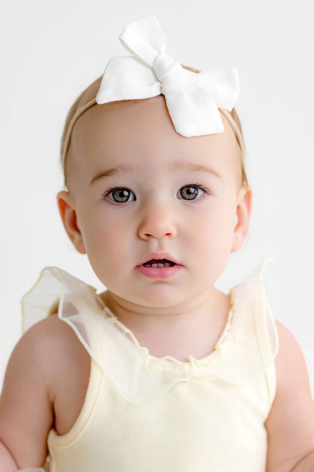 Adorable baby girl wearing a white bow headband and yellow ruffle romper, looking at camera with big blue eyes.