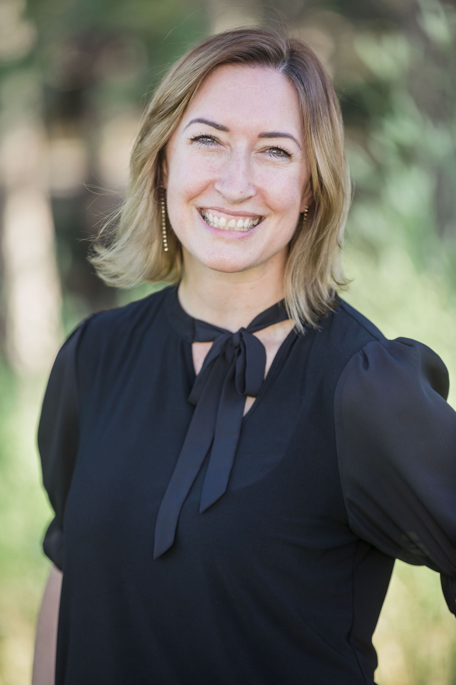 Smiling woman with blonde hair wearing a black tie-neck blouse, posing outdoors with trees in background.