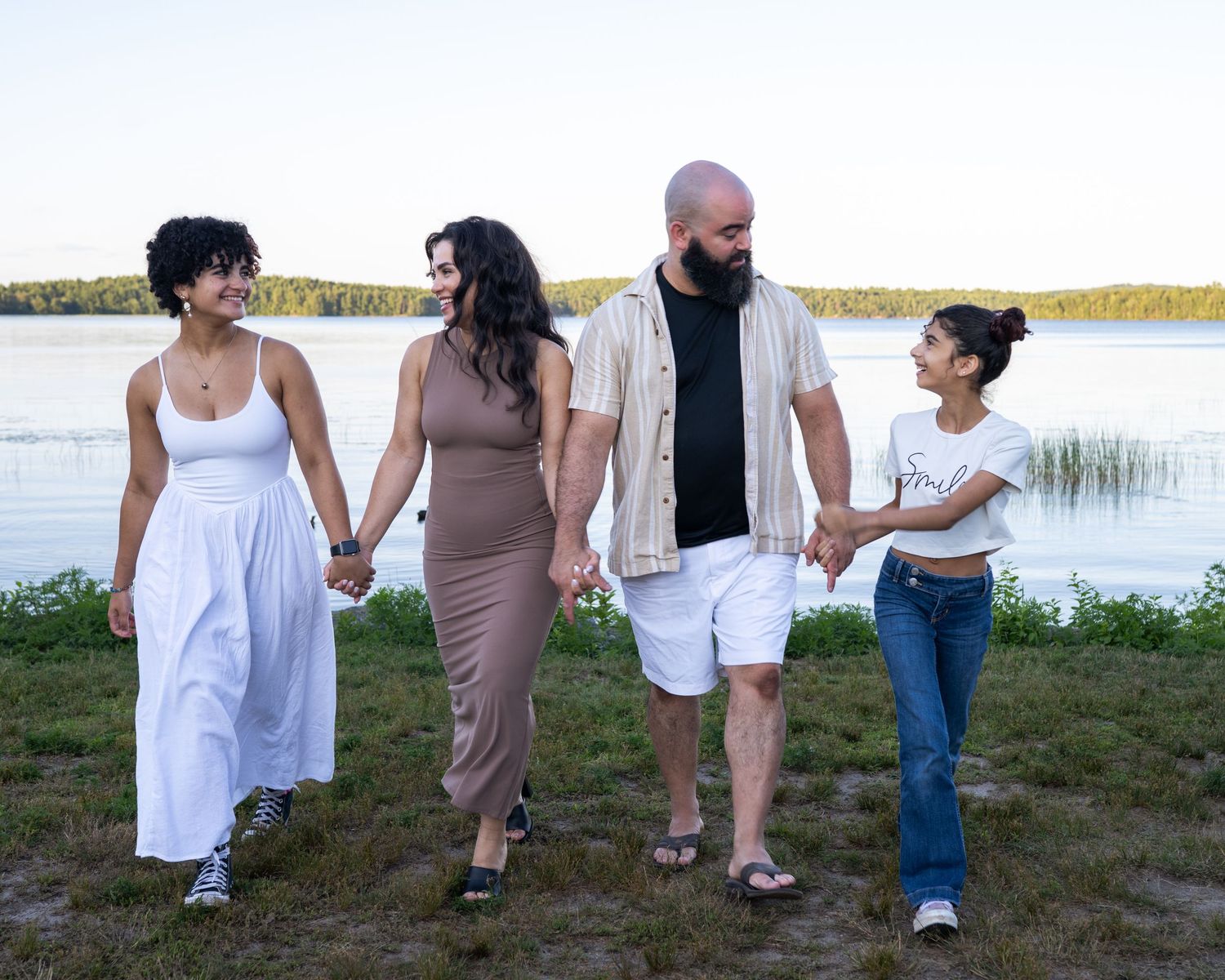 Family of four holding hands walking near a serene lake at sunset on a grassy shore.