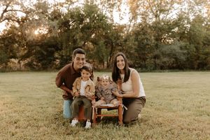 Family of four smiling outdoors at golden hour, parents kneeling beside two young children on a small bench in a green field.