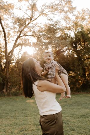 A mother lifts her smiling baby girl in a sunlit park during golden hour, surrounded by lush green trees.