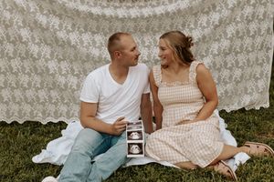Couple sitting on grass holding ultrasound photos during outdoor pregnancy announcement with lace backdrop.