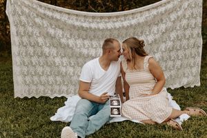 Couple sharing a tender moment on a blanket with ultrasound photos, in front of a lace backdrop outdoors.