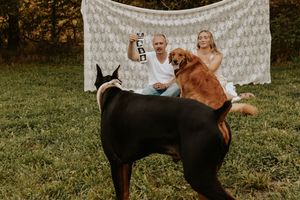 Couple sitting outdoors with two dogs, a Rottweiler and Golden Retriever, in front of a white lace backdrop.