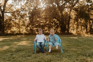 Two young boys sitting together in blue chairs on a sunny green lawn surrounded by golden trees.