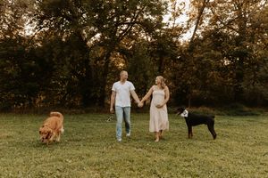 Pregnant couple holding hands walking with golden retriever and black dog in a lush green park at golden hour.