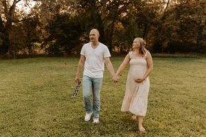 Pregnant couple holding hands and walking in a green field surrounded by trees during golden hour.