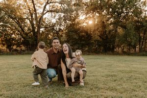 Happy family of four posing outdoors at golden hour in a lush green park, parents laughing with two young toddlers.