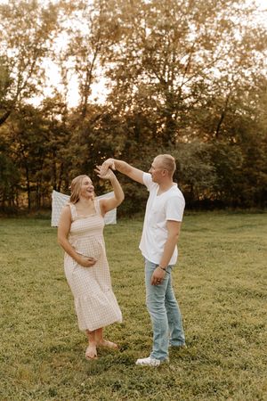A couple dances outdoors in a field, the woman in a pink dress cradling her baby bump during a maternity photo session.