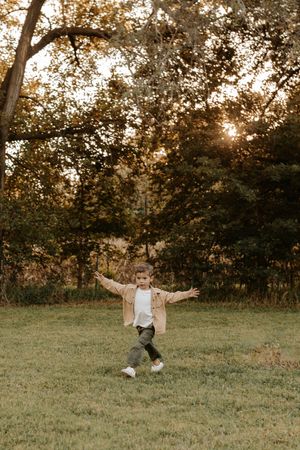 Young boy running outdoors in autumn park, wearing beige jacket, arms outstretched, golden sunset light filtering through trees.