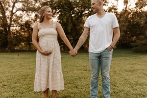 Pregnant woman in gingham dress holding hands with man in white tee on a green lawn outdoors.