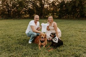 Couple kneeling on grass holding baby onesie with two dogs wearing Big Bro bandanas for pregnancy announcement.