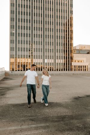 Couple holding hands walking on rooftop parking lot with modern glass office building in urban city background.