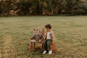 Two young children sit together on a wooden chair in a sunny green field surrounded by trees.