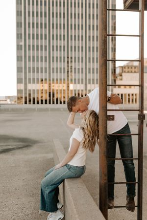 Couple sharing a kiss on a rooftop parking garage with city buildings in the background, both wearing white tees and jeans.