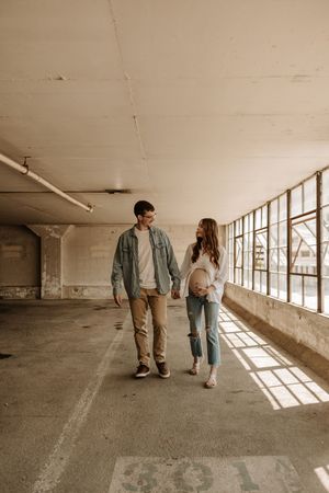 Couple holding hands walking through an urban parking garage with natural light streaming through windows.