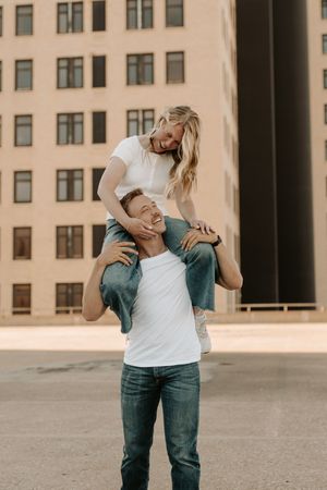A man gives a laughing blonde woman a piggyback ride outdoors in front of a modern building.