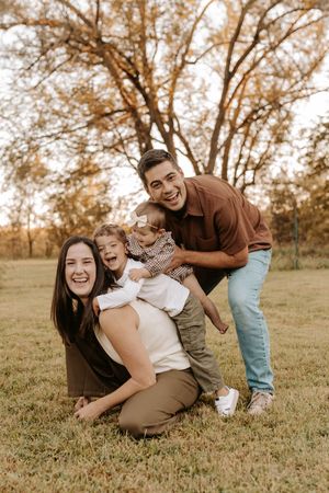 Happy family of four poses outdoors in autumn park, parents smiling with two young children on green grass.