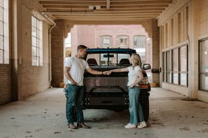 A couple stands facing each other beside a black truck inside an industrial building with brick walls.