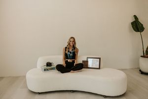Smiling blonde woman sitting cross-legged on a white curved sofa in a minimalist room with a plant and books nearby.