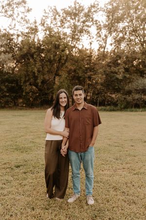 A couple smiles while holding hands in a grassy park, woman in white top and olive pants, man in brown shirt and jeans.