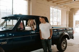 Couple posing with a vintage blue Ford F-150 truck inside an industrial building, woman seated inside, man leaning on door.