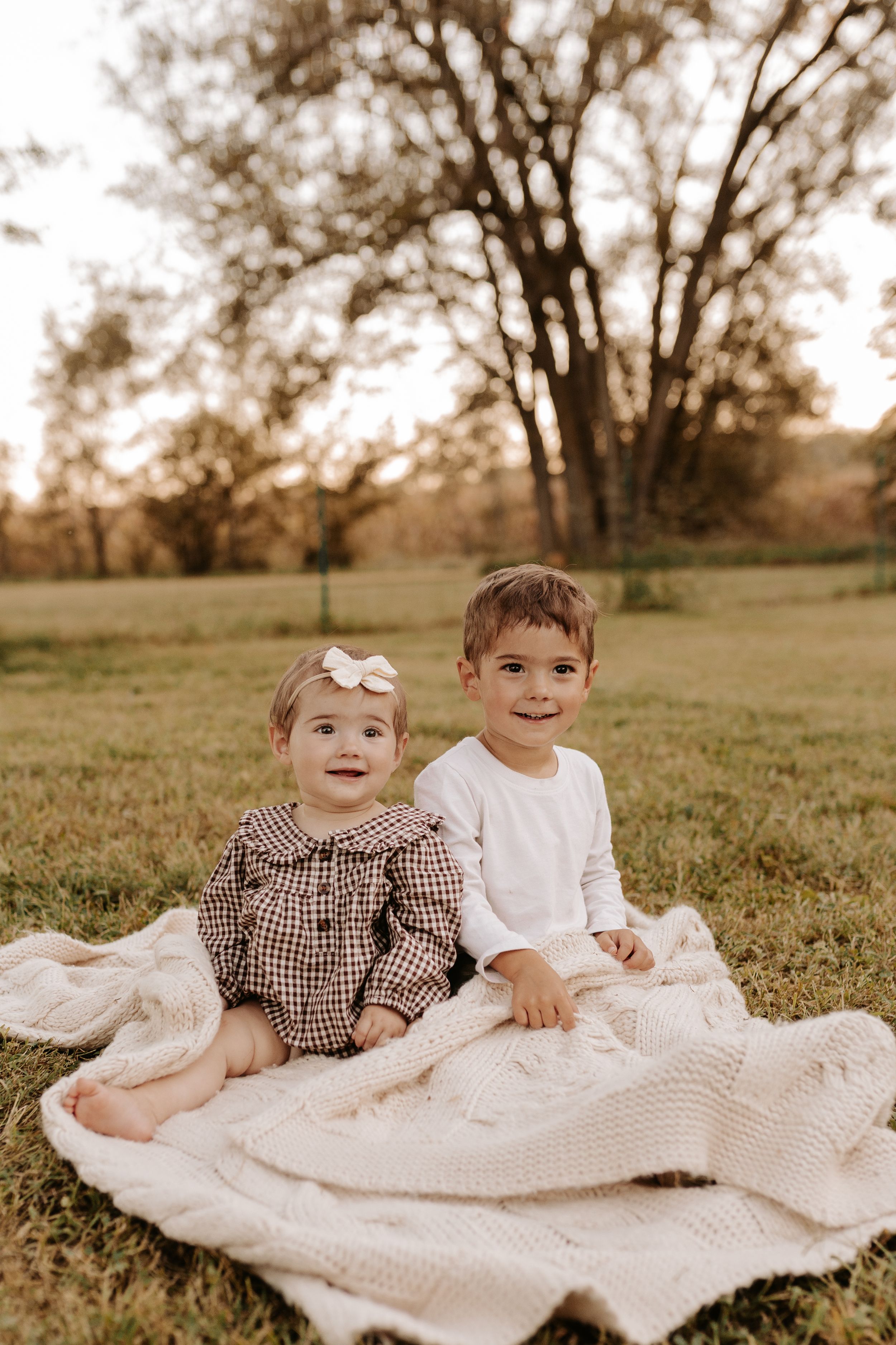 Two young siblings sit smiling on a cream knit blanket outdoors at golden hour, girl in plaid dress with bow, boy in white.