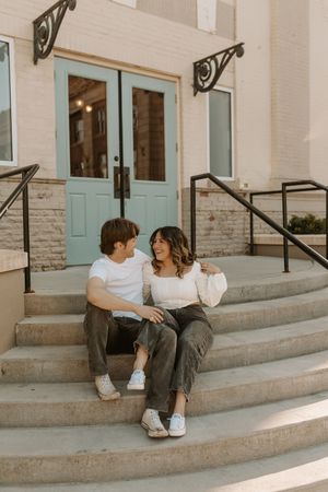 A couple sits together on stone steps outside a building with teal doors, smiling at each other in casual white tops and olive pants.