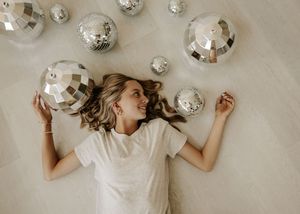 Woman lying on floor surrounded by silver disco balls, holding one and smiling, wearing a white t-shirt.
