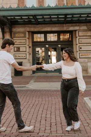 A joyful couple holds hands and dances on a brick plaza outside a historic station building.