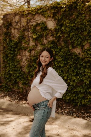 Smiling pregnant woman in white shirt and jeans posing outdoors against ivy-covered stone wall.