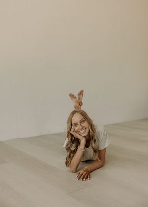 Smiling young woman with wavy blonde hair lying on floor in white tee, resting chin on hand in minimalist studio.